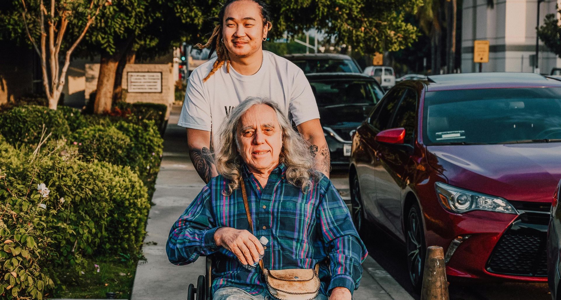 A caring scene of a young man pushing a senior in a wheelchair along a sidewalk.