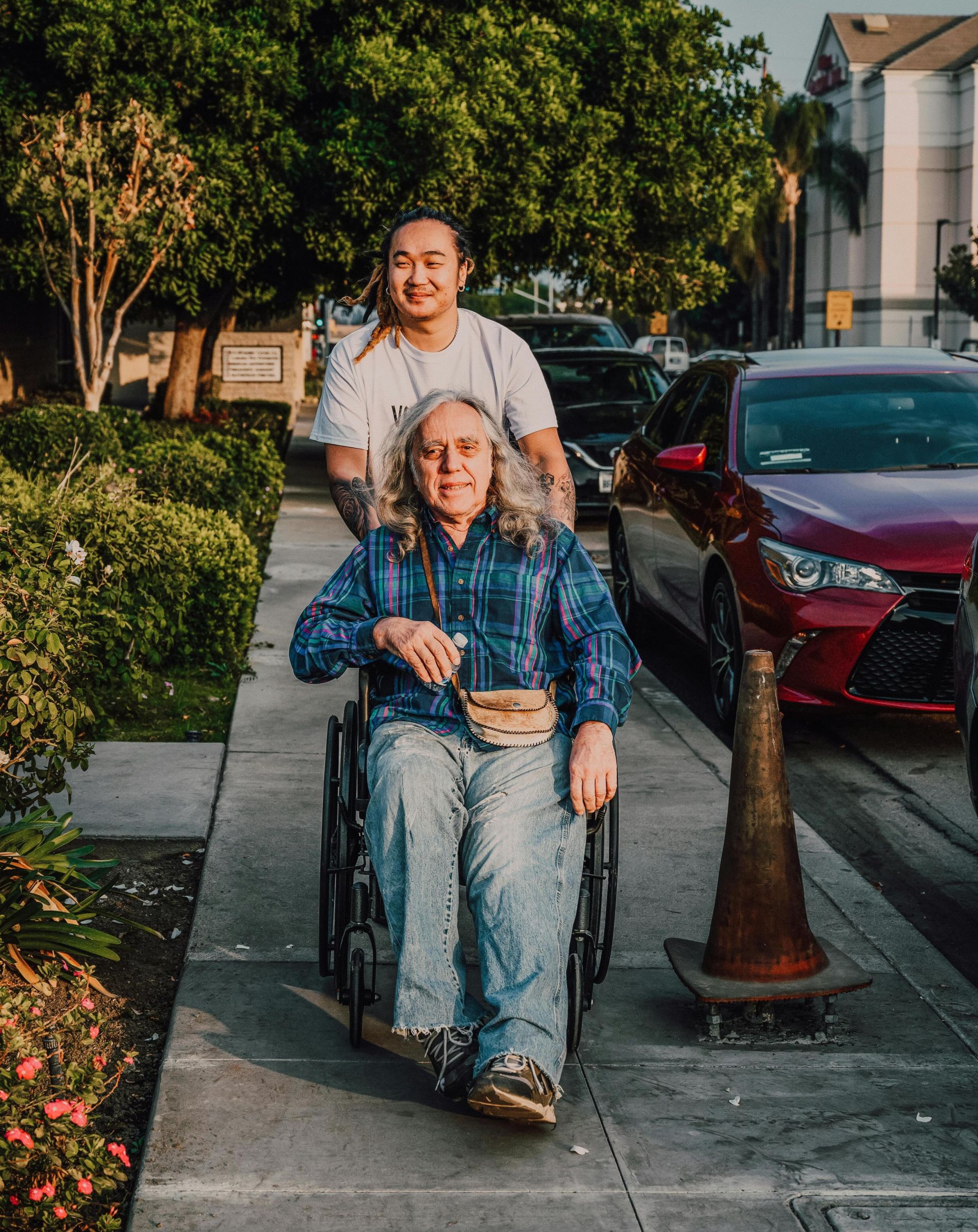 A caring scene of a young man pushing a senior in a wheelchair along a sidewalk.